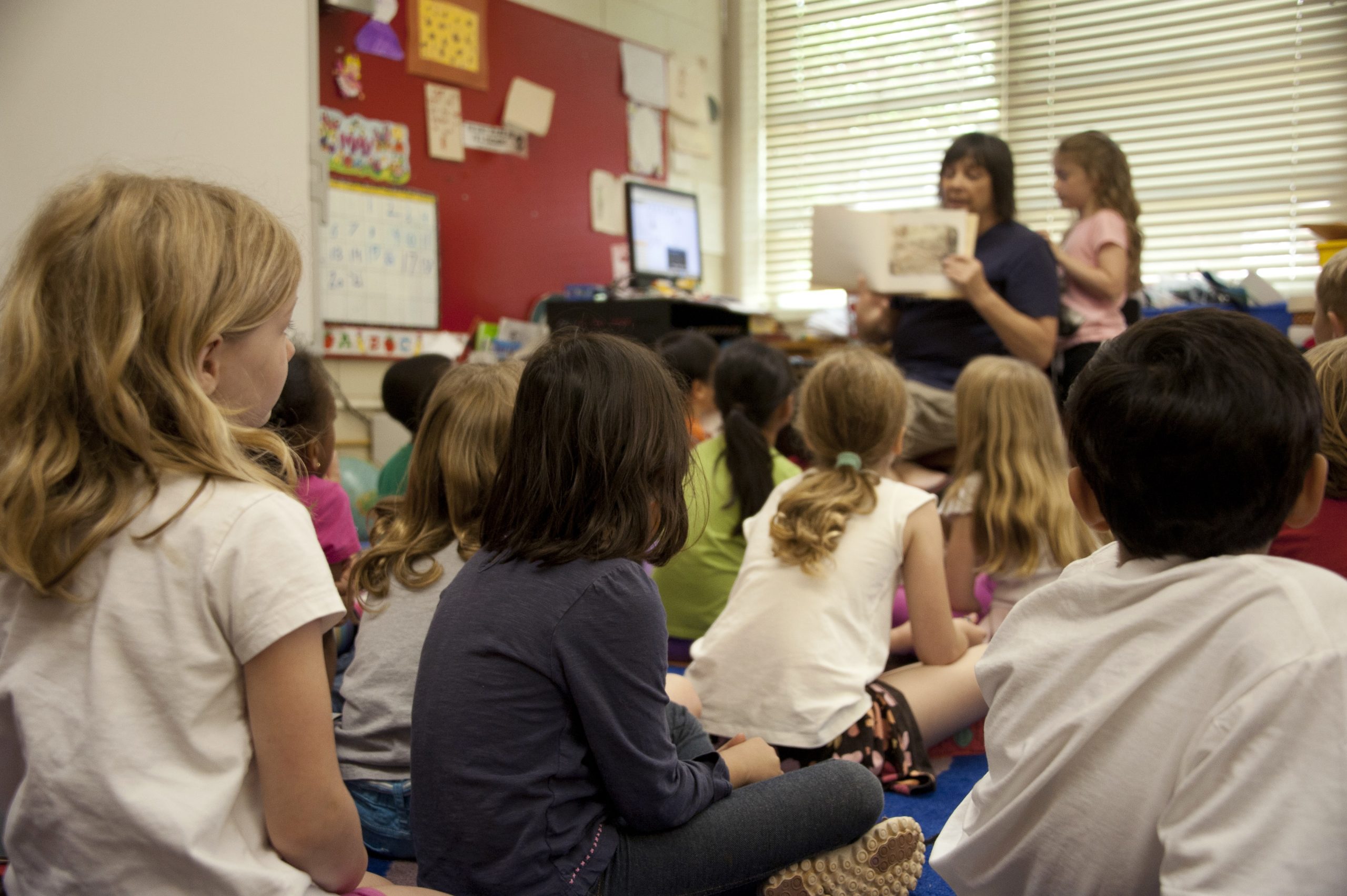 Foto de alumnado en clase con maestra al fondo leyendo